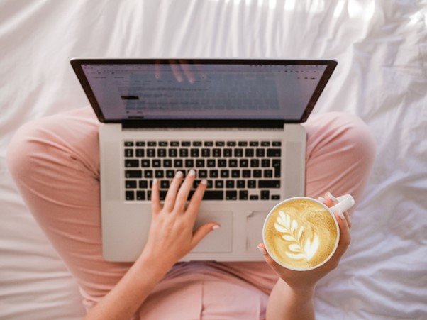 Woman working remotely on laptop in bed holding coffee