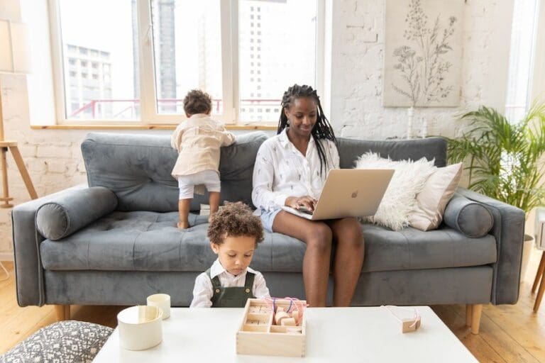 Mother remote working on laptop on sofa whilst kids play around her