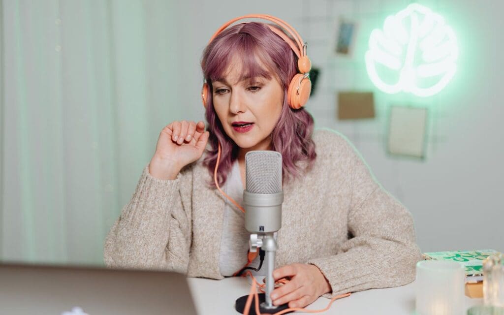Woman with pink hair wearing peach coloured headphones recording a podcast with a microphone and a MacBook