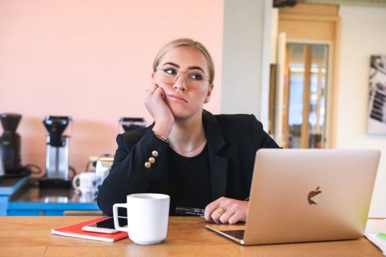 Woman looking fed-up at her desk in front of a Macbook and a cup of coffee, burnout concept