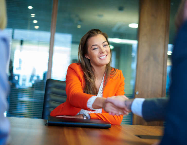 Woman with brown hair wearing an orange blazer in a job interview, shaking hands with the interviewer and smiling