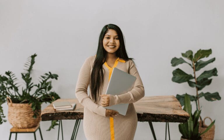 Female founder standing in front of a wooden desk holding a laptop surrounded by green plants