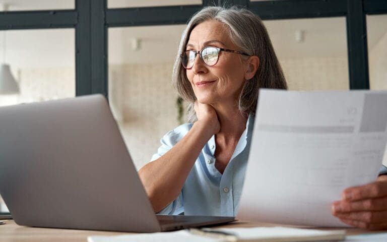 Over-50s woman in tech looking out into the distance with a laptop on her desk and a piece of paper in her hand