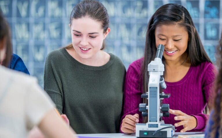 Teenage girls in a classroom with one looking into a scientific microscope, girls in STEM concept