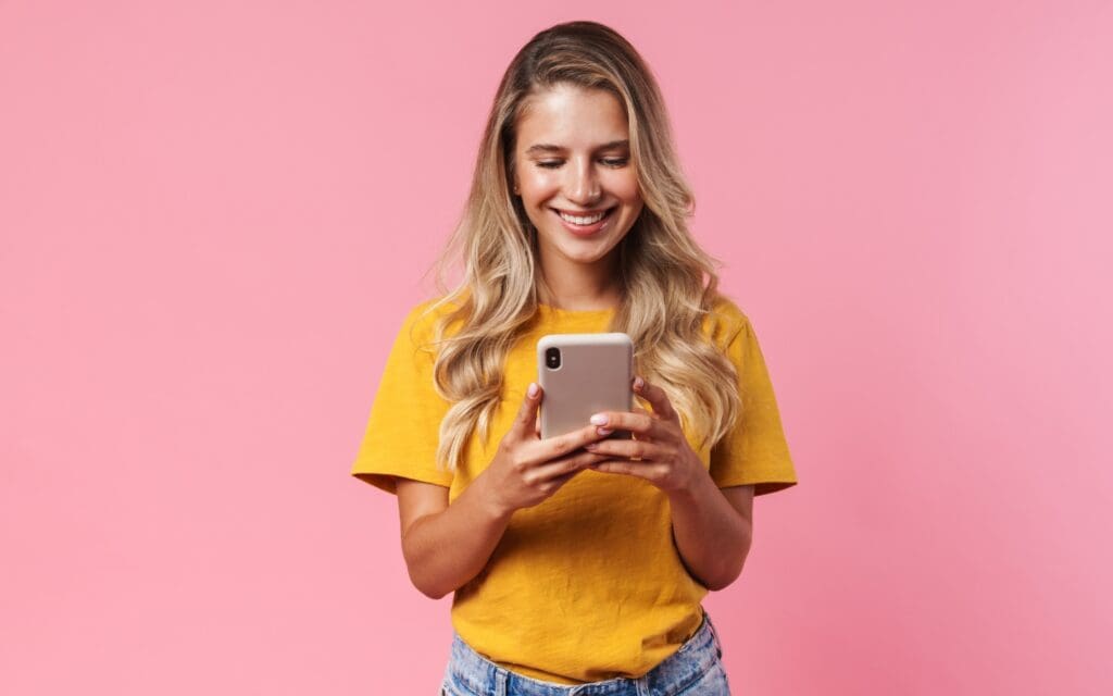 Woman wearing a yellow t-shirt texting on a pink phone in front of a pink background
