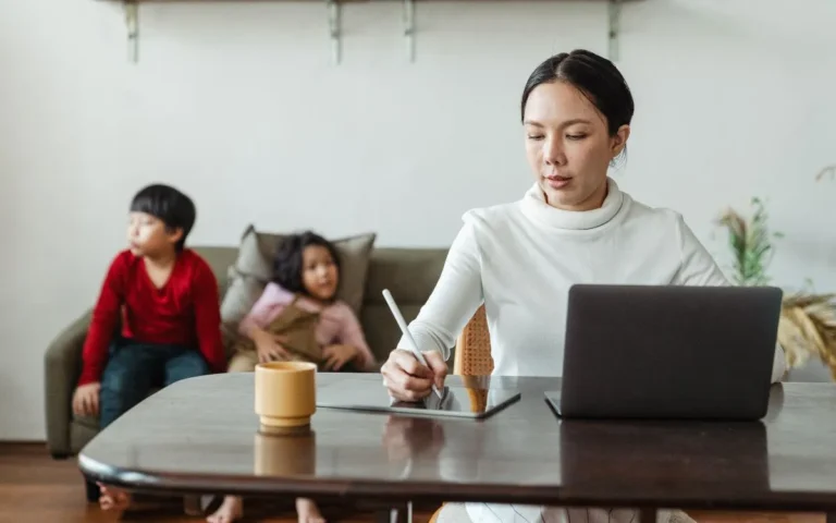 Mum working on laptop at table with bored children behind, working mum concept