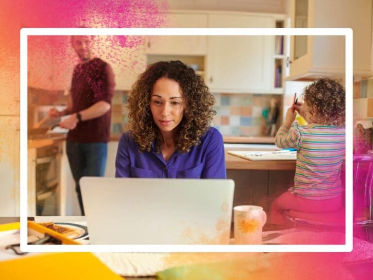 A working mum sitting at the dining table typing on a laptop, while her child draws behind her and partner cooks breakfast