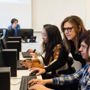 College students coding with teacher in tech lab classroom.